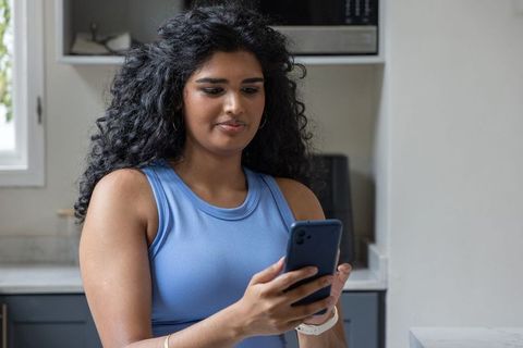 Indian woman using smartphone in modern kitchen wearing blue sleeveless top, scrolling