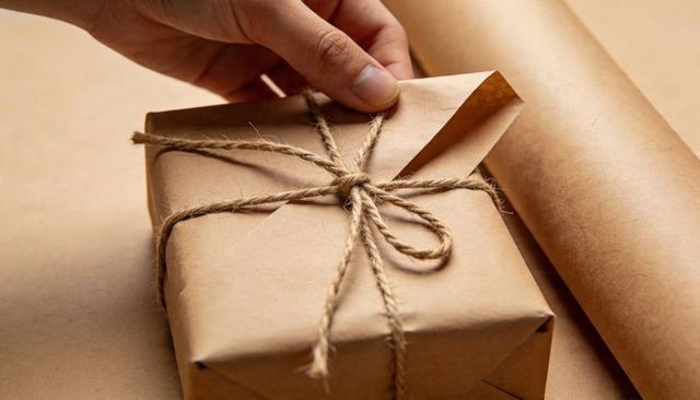 Lifting kraft paper corner from twine-wrapped gift box on craft table minimalist styling
