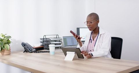 African woman doctor using tablet and phone in minimalist clinic office with digital tools