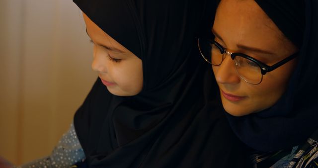 Mother and daughter in hijabs spending quality time reading indoors. Useful for topics on Muslim families, education, cultural practices, family bonding, parental support, and the role of mothers in upbringing.