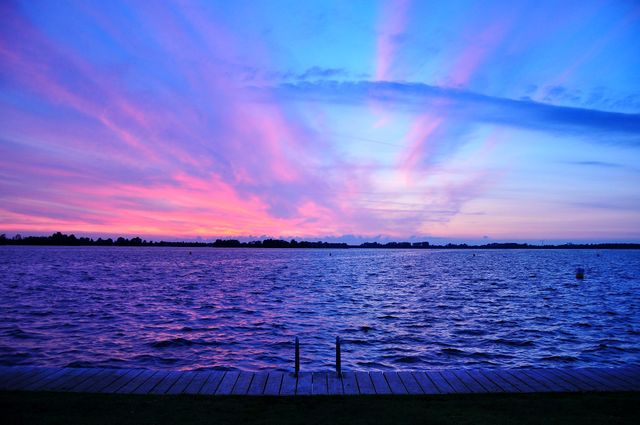 Stunning sunset over a tranquil lake with vibrant hues of pink, purple, and blue dominating the sky. The calm water reflects the colorful sky, and a wooden pier leads into the lake. Perfect for backgrounds, travel blogs, nature calendars, or inspirational posters.