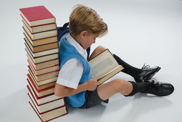Young boy in school uniform reading a book while sitting against a tall stack of books on a white background. Ideal for educational materials, school promotions, literacy campaigns, and children's book advertisements.