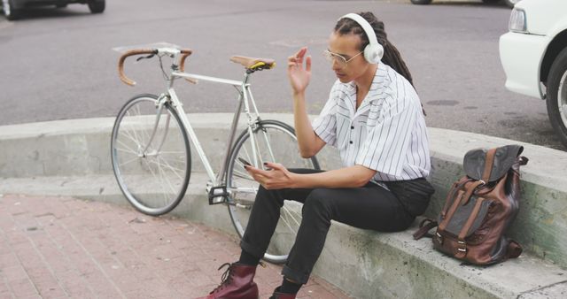 Side view of a biracial man with long dreadlocks out and about in the city on a sunny day, sitting in the street, wearing headphones, using a smartphone, with his bicycle standing next to him in slow motion.
