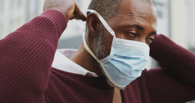 This image features a man wearing a protective face mask in an urban area. He is adjusting the mask, demonstrating caution and awareness of public health measures. Suitable for use in topics related to health safety, COVID-19 precautions, and urban lifestyle in the new normal. Ideal for articles, health campaigns, and informational material on protective measures.