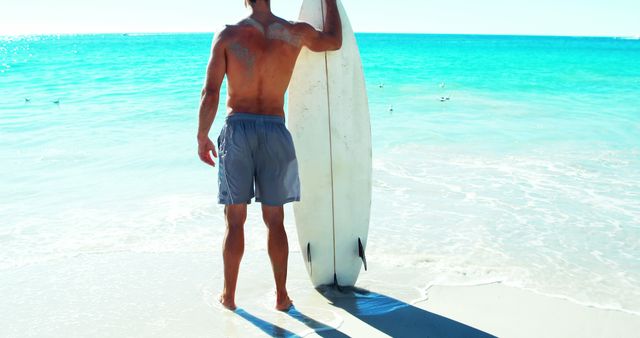 Man stands on shore holding his surfboard, looking out to the turquoise ocean waves. Ideal for travel brochures, surf-related businesses, summer ads, and beach lifestyle promotions.