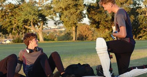 Teenage cricket teammates laughing and strapping batting pads on sunlit field