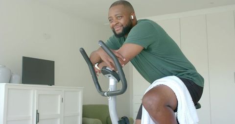 Smiling african man leaning on exercise bike in bright modern living room wearing earbuds