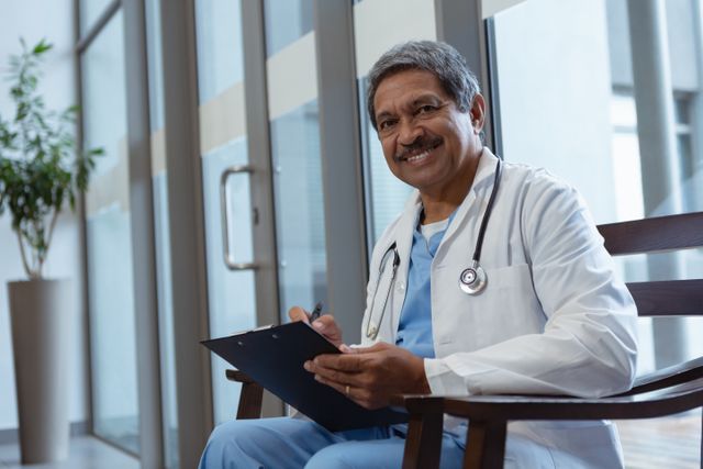 Mature male doctor sitting in hospital lobby, writing on clipboard. Ideal for healthcare, medical, and professional themes. Useful for illustrating patient care, medical consultations, and healthcare environments.