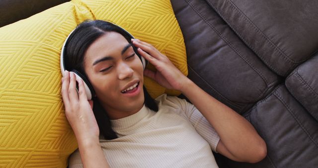 Gender fluid male wearing headphones enjoying listening to music while lying on the couch at home. concept of gender expression, identity and diversity