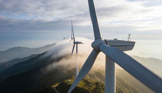 Wind turbine nacelle and blades on foggy mountain ridge with winding access road at sunrise