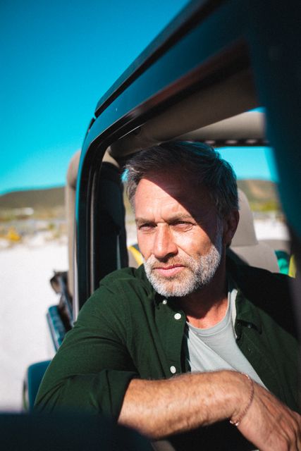 Mature caucasian man with a beard sitting in an off-road vehicle, looking away thoughtfully during a summer road trip. Ideal for use in travel blogs, vacation advertisements, adventure magazines, and lifestyle websites promoting outdoor activities and road trips.