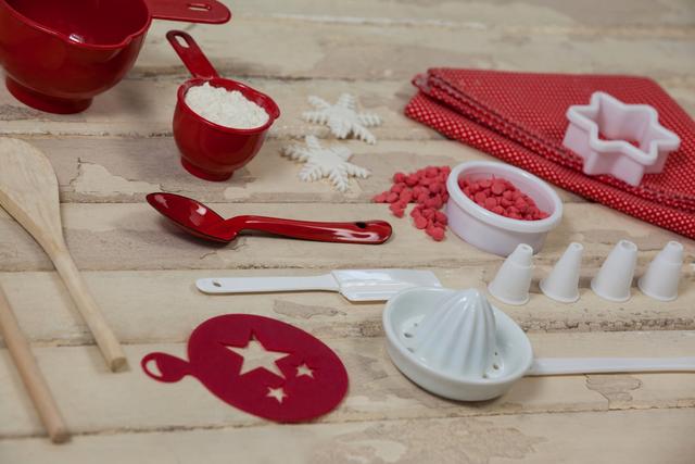 A variety of baking utensils and ingredients arranged on a wooden board. Red and white measuring spoons, a wooden spatula, flour, and decorative toppings are displayed. Ideal for use in food blogs, recipe books, and kitchenware advertisements.