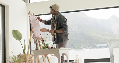 Indian artist painting on easel in sunlit studio overlooking waterfront and mountain