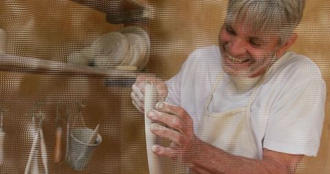 Senior potter shaping tall ceramic vessel on wheel wearing beige apron and smiling