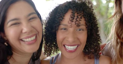 Diverse female friends smiling and laughing while taking selfie in sunlit park with backlit bokeh