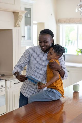 African american father hugging son while reading together on kitchen counter, cozy home