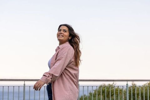 Indian woman smiling by ocean railing wearing pink overshirt and denim casual portrait