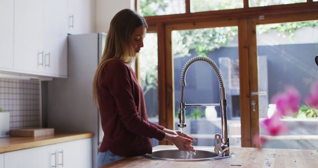 Young woman practicing good hygiene washing hands in kitchen. Large window providing natural light, modern appliances, and wooden countertops suggest a contemporary and clean living space. Useful for lifestyle, health, and domestic life topics.
