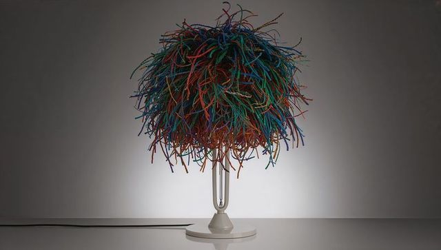 Multicolor pom-pom table lamp casting soft halo on gray wall, glossy white base, tangled shade