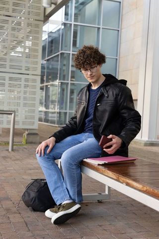 Curly teen male sitting on plaza bench checking smartphone with notebooks