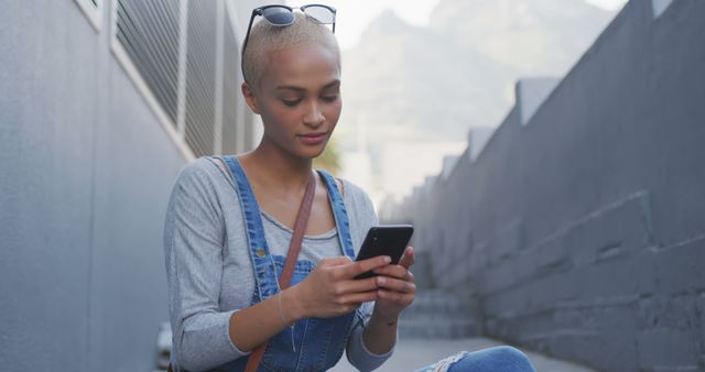 Young woman with short hair and sunglasses on her head using a smartphone. She is sitting outdoors in an urban setting, likely texting or browsing. Can be used for themes related to technology, communication, casual lifestyle, or modern city life.
