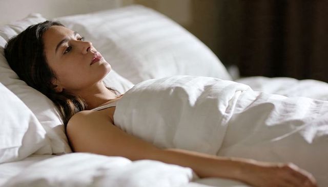 Young woman lying in bed gazing upward, resting under white duvet, serene bedroom moment