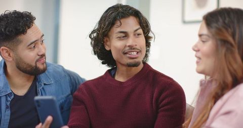 Multiracial friends sharing smartphone on couch during casual living room conversation