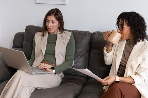 Diverse female coworkers reviewing laptop and documents while sipping coffee on sofa