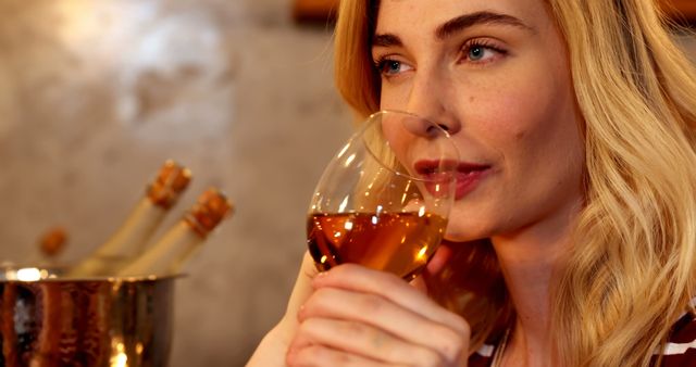 Blonde woman in cozy atmosphere savoring a glass of white wine. Her serene expression and the soft indoor lighting create a relaxed and elegant mood, ideal for use in content related to leisure, lifestyle, wine tasting events, relaxation, and sophisticated evenings.