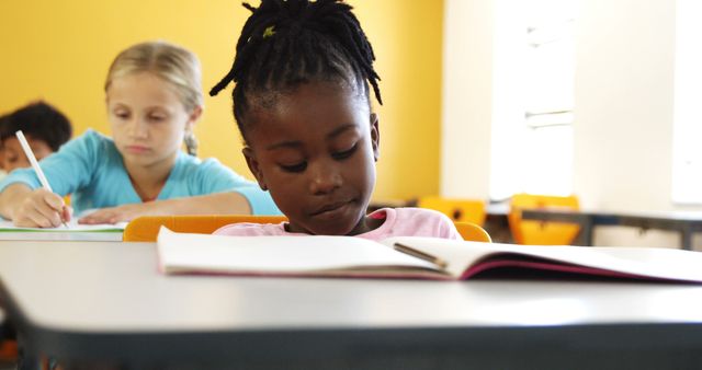 School children are diligently studying in a classroom setting. The primary focus is an African American girl deeply engaged in reading her book, while a Caucasian girl in the background is writing in her notebook. This can be used for educational content, diversity awareness, school promotions, and teaching resources.