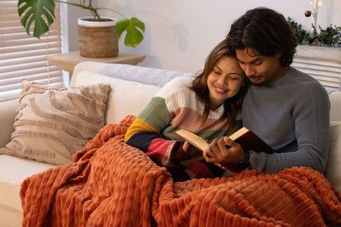 Diverse couple cuddling and reading on cozy sofa with rust blanket and textured pillows