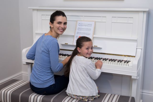 Mother and daughter sitting together at a white piano, with sheet music in front of them. Both are smiling and looking at the camera, creating a warm and joyful atmosphere. Ideal for use in family-oriented content, music education materials, and advertisements promoting family bonding and learning activities.