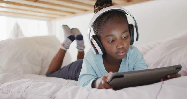 Happy african american girl in headphones using tablet lying on bed at home, copy space. Technology, communication and domestic life.