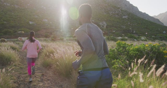 Two people jogging on a nature trail surrounded by mountains and vegetation, with sunlight shining through. Perfect for advertising fitness routines, outdoor activities, wellness programs, and health-conscious lifestyles.