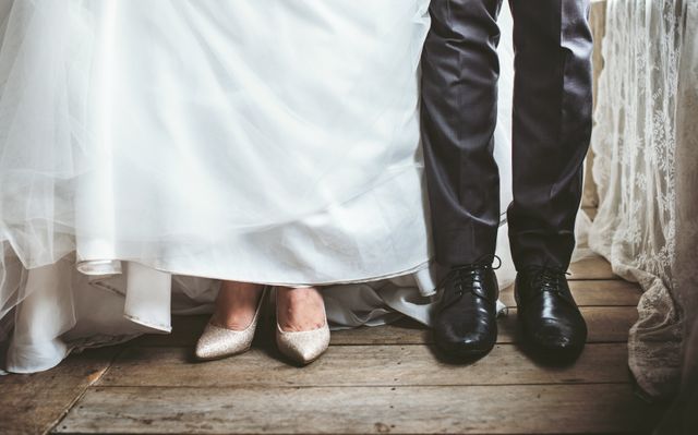 Captures just the feet of the bride and groom on their wedding day. The image highlights the contrast between the bride's elegant shoes and the groom's polished black shoes, set against a rustic wooden floor. Ideal for wedding invitations, blogs on wedding fashion, and matrimonial websites.