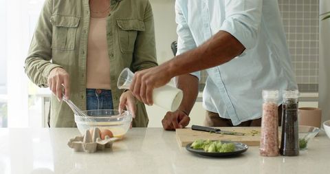 Couple whisking eggs and pouring milk at bright kitchen island for homemade breakfast