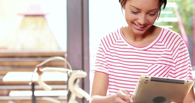A young African American woman is smiling while using a tablet, with copy space. Her casual attire and the cozy indoor setting suggest she could be enjoying leisure time or engaging in remote work.