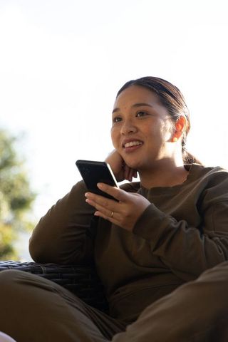 Sunlit asian woman relaxing on patio holding smartphone and smiling in olive loungewear