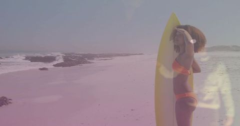 Surfer standing with yellow surfboard shielding eyes on sunny sandy beach with copy space