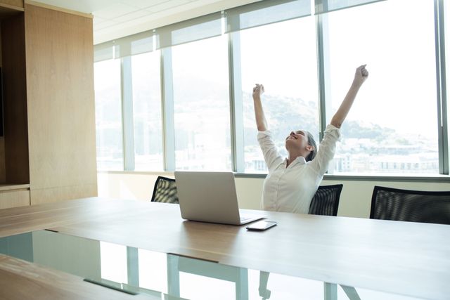 Businesswoman sitting in a modern conference room with arms raised in celebration. Ideal for illustrating success, achievement, and motivation in a professional setting. Suitable for business, corporate, and workplace-related content.