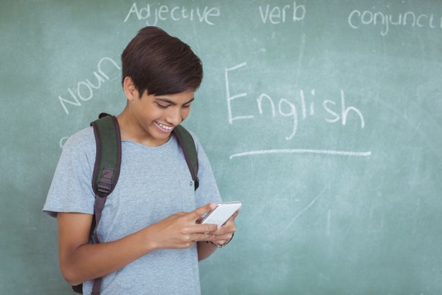 A schoolboy with a backpack is using a mobile phone in a classroom. The chalkboard behind him has English words written on it. This image can be used for educational content, technology in education, school-related advertisements, or articles about student life and learning.