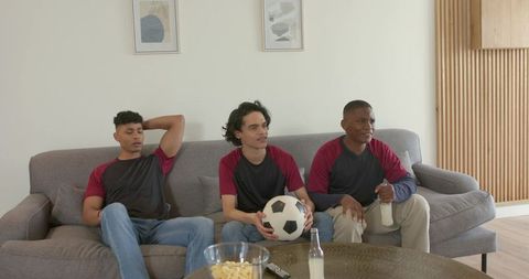 Diverse male friends watching soccer on couch holding ball and drinks relaxing together