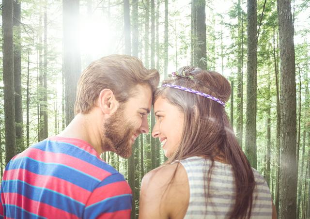 Couple standing in forest with tall trees and sunlight filtering through. They are facing each other and smiling, forehead to forehead, suggesting moments of bonding and affection. The image captures a serene and romantic outdoor scene, making it suitable for uses in nature-related projects, romantic themes, relationship articles, and lifestyle blogs.