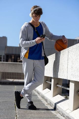 Teen athlete leaning on urban rooftop parapet checking phone and holding basketball