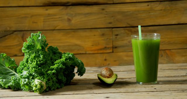 A bunch of fresh kale and a halved avocado are placed next to a glass of green smoothie on a wooden table, with copy space. These ingredients suggest a focus on healthy eating and the preparation of a nutritious vegetable-based drink.