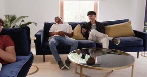 Multiracial Young Men Relaxing on Navy Velvet Sofa, One Smiling While Using Smartphone