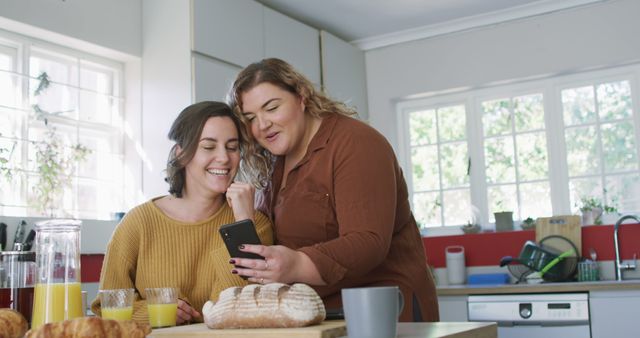 Two women are enjoying a relaxed moment in a modern kitchen, sharing a laugh while looking at a smartphone. There is a table with breakfast items such as a loaf of bread, croissants, and juice in front of them. This scene is perfect for depicting friendship, casual home moments, and the joy of everyday life. It can be used for advertising home products, lifestyle blogs, or promotions related to technology and social media.