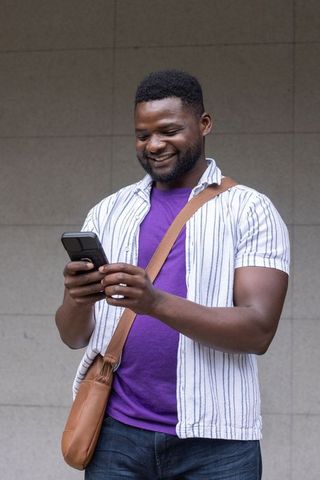 Smiling african american man using smartphone wearing purple tee and brown crossbody bag