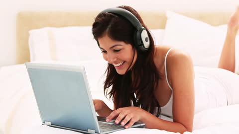 Smiling Woman Using Laptop with Headphones Lying on Bed