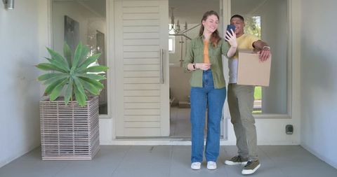 Young couple smiling and taking selfie at new home doorway while holding moving box